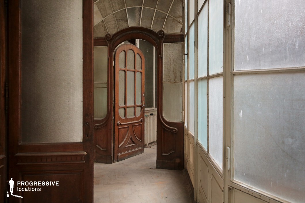 This image features an elegant yet aged interior hallway with intricate wooden door frames and frosted glass panels, evoking an early 20th-century aesthetic. The central arched door, adorned with curved decorative elements, stands partially open, leading to another softly lit space. The surrounding walls feature textured, semi-transparent glass, allowing diffused light to enter and enhance the nostalgic atmosphere. The worn parquet floor and subtle architectural details add to the sense of history. This location is ideal for period dramas, psychological thrillers, or mystery films, offering an evocative setting for quiet revelations, eerie encounters, or scenes of hidden secrets.