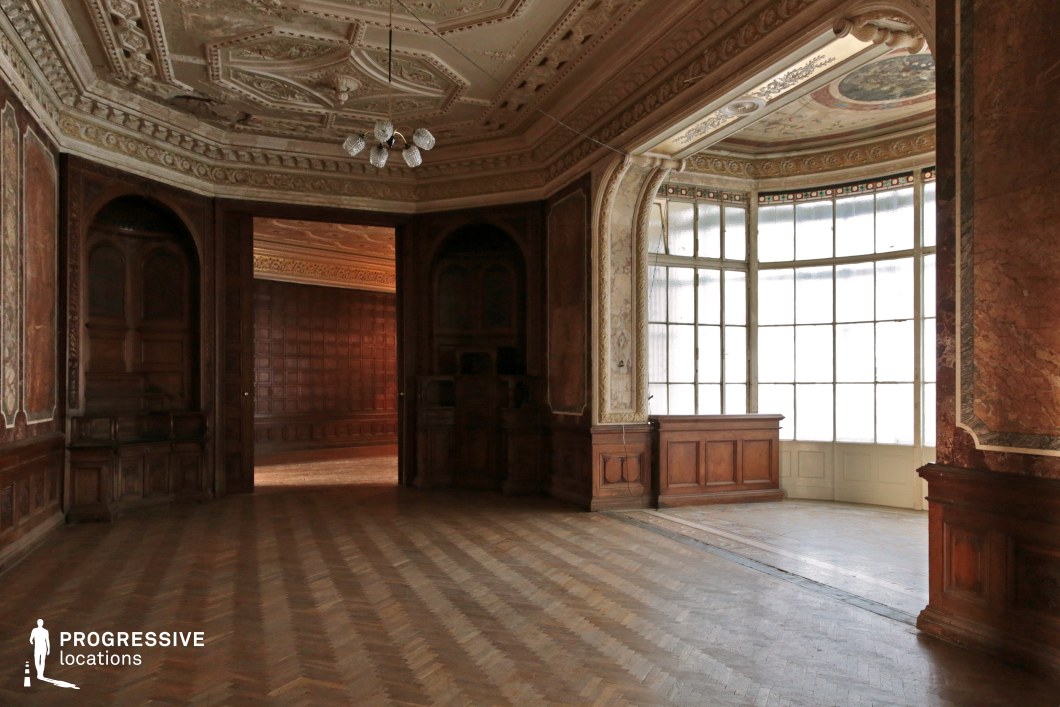 This image showcases a grand yet slightly worn ballroom with elaborate ceiling moldings, richly detailed wooden paneling, and a spacious layout. The large bay window, framed by ornate arches, allows soft natural light to filter in, illuminating the parquet flooring and decorative wall frescoes. The adjoining room, visible through a wide doorway, features dark wood paneling and a stately ambiance, adding depth and continuity to the space. The mix of opulence and subtle decay makes this location ideal for historical dramas, aristocratic period films, gothic mysteries, or psychological thrillers, offering an atmospheric and visually rich backdrop for cinematic storytelling.
