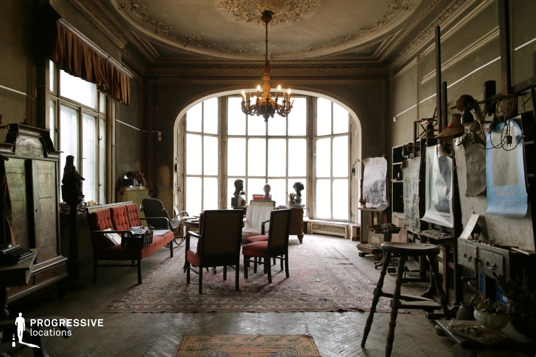 This image showcases a richly detailed, timeworn studio space with an arched bay window that fills the room with soft, diffused light. The ornate ceiling moldings, antique furnishings, and worn wooden floors create an atmosphere of artistic nostalgia. The presence of easels, framed artworks, and decorative busts suggests a creative or intellectual setting, while the warm tones of the upholstered chairs and patterned rugs contrast with the dark patina of the walls. This location is ideal for filming period dramas, historical mysteries, artistic biopics, or psychological thrillers, offering a visually textured and evocative setting with a sense of character and history.