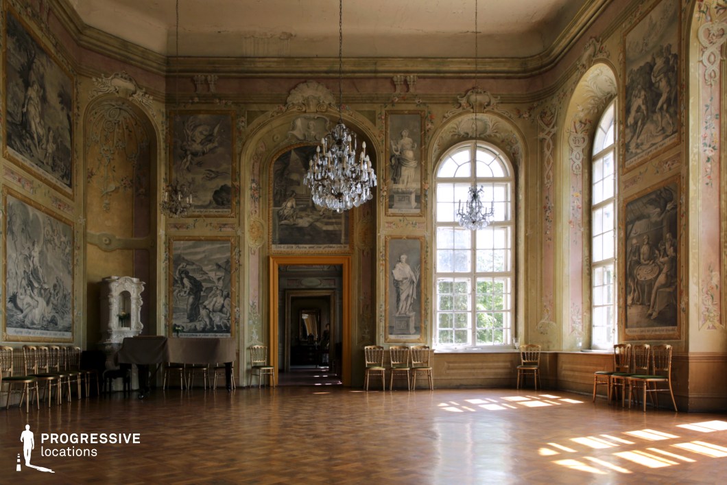 This image showcases a grand ballroom adorned with intricate frescoes, high arched windows, and shimmering chandeliers. The detailed wall paintings, framed by delicate stucco moldings, depict classical scenes, enhancing the space’s historical elegance. The polished parquet flooring reflects the natural light streaming in through the large windows, which offer glimpses of lush greenery outside. The room's symmetry, ornate decor, and timeless grandeur make it an ideal location for filming period dramas, historical romance, aristocratic intrigue, or fantasy narratives, providing a setting rich in visual depth and refined sophistication.