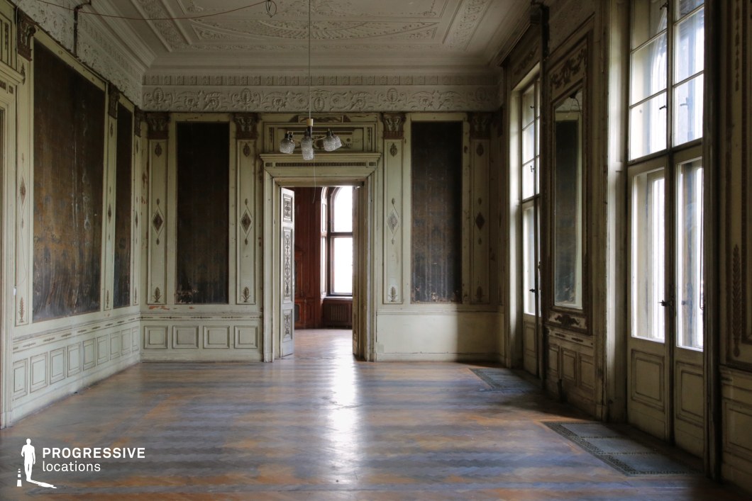 This image captures an opulent yet decayed neoclassical-style room, featuring high ceilings with intricate plasterwork, tall windows, and elaborately paneled walls. The faded and peeling wall decorations hint at former grandeur, while the worn parquet flooring adds to the room’s aged character. A set of double doors opens into an adjoining space, framed by carved moldings and a glimpse of richly toned wooden paneling beyond. The large windows allow natural light to filter in, enhancing the atmospheric contrast between elegance and neglect. This location is ideal for filming period dramas, psychological thrillers, or mystery films, providing a compelling backdrop for narratives of lost luxury, hidden secrets, or aristocratic decline.