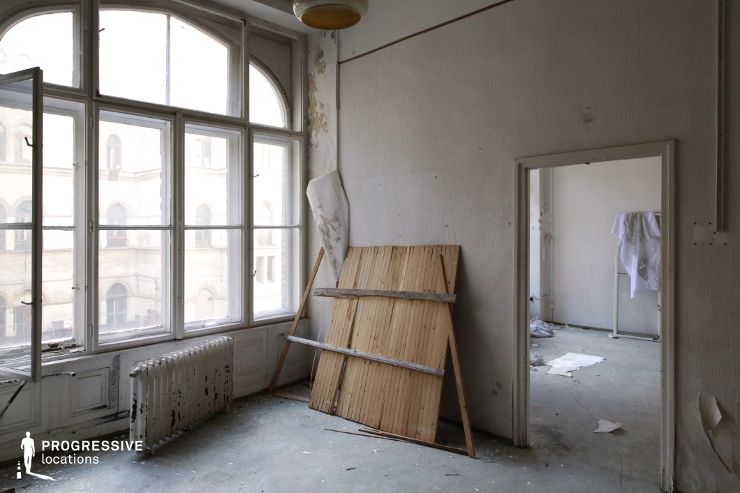 This deteriorating interior features expansive arched windows that flood the space with soft natural light, contrasting with the peeling paint, cracked walls, and scattered debris. The aged wooden radiator and exposed framework add to the atmosphere of neglect, while the adjoining room, partially visible through an open doorway, deepens the sense of emptiness. The muted color palette of whites and grays, combined with the worn textures, creates a melancholic yet visually striking scene. This location is ideal for filming psychological dramas, art-house films, or dystopian narratives, providing a setting that conveys themes of isolation, forgotten history, and quiet decay.