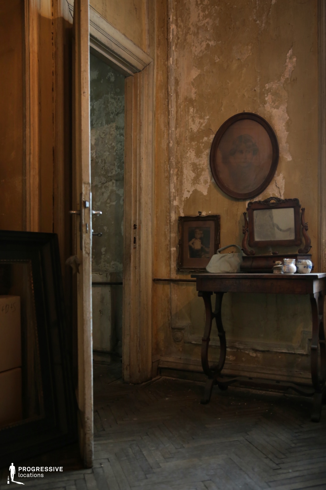 This image captures a dimly lit, timeworn interior with peeling walls, faded paint, and antique furnishings. The delicate wooden table, adorned with a small mirror and framed photographs, adds to the nostalgic and intimate atmosphere. The ajar door leads into another shadowy space, enhancing the sense of depth and mystery. The herringbone-patterned wooden flooring, aged picture frames, and worn textures evoke a bygone era. This location is ideal for psychological thrillers, historical dramas, gothic horror, or period films, offering a moody and visually layered setting rich in history and emotion.