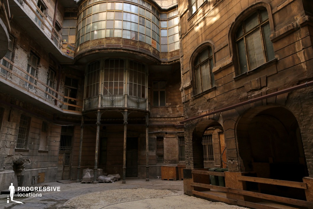 This image presents a worn, enclosed courtyard surrounded by aged buildings with peeling facades and darkened stone walls. The most striking feature is a curved glass bay window, supported by delicate iron columns, which adds an air of faded elegance. The structure shows visible decay, with crumbling plaster, broken masonry, and rusted metal railings accentuating its abandoned or neglected state. The arched doorways and dim passageways create a sense of depth and mystery, making this an ideal location for filming atmospheric historical dramas, noir thrillers, dystopian narratives, or crime films that require a moody, immersive setting.
