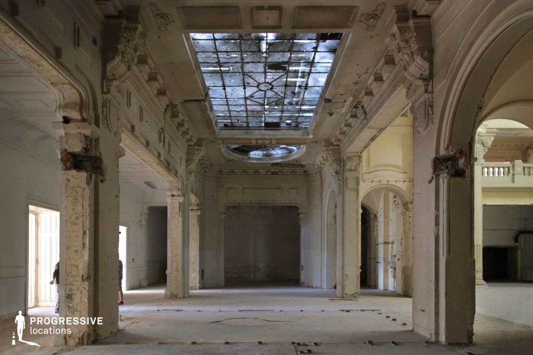 This image showcases a vast, decayed hall with intricate classical detailing and a partially collapsed skylight. The tall arches, sculpted moldings, and faded white surfaces evoke a sense of former grandeur now overtaken by time. The broken glass ceiling allows diffused natural light to filter through, casting a soft glow on the worn floors and crumbling walls. The expansive space and high ceilings create an imposing atmosphere, ideal for filming historical dramas, dystopian narratives, psychological thrillers, or mystery films, where themes of abandonment, lost opulence, or hidden secrets are central to the visual storytelling.