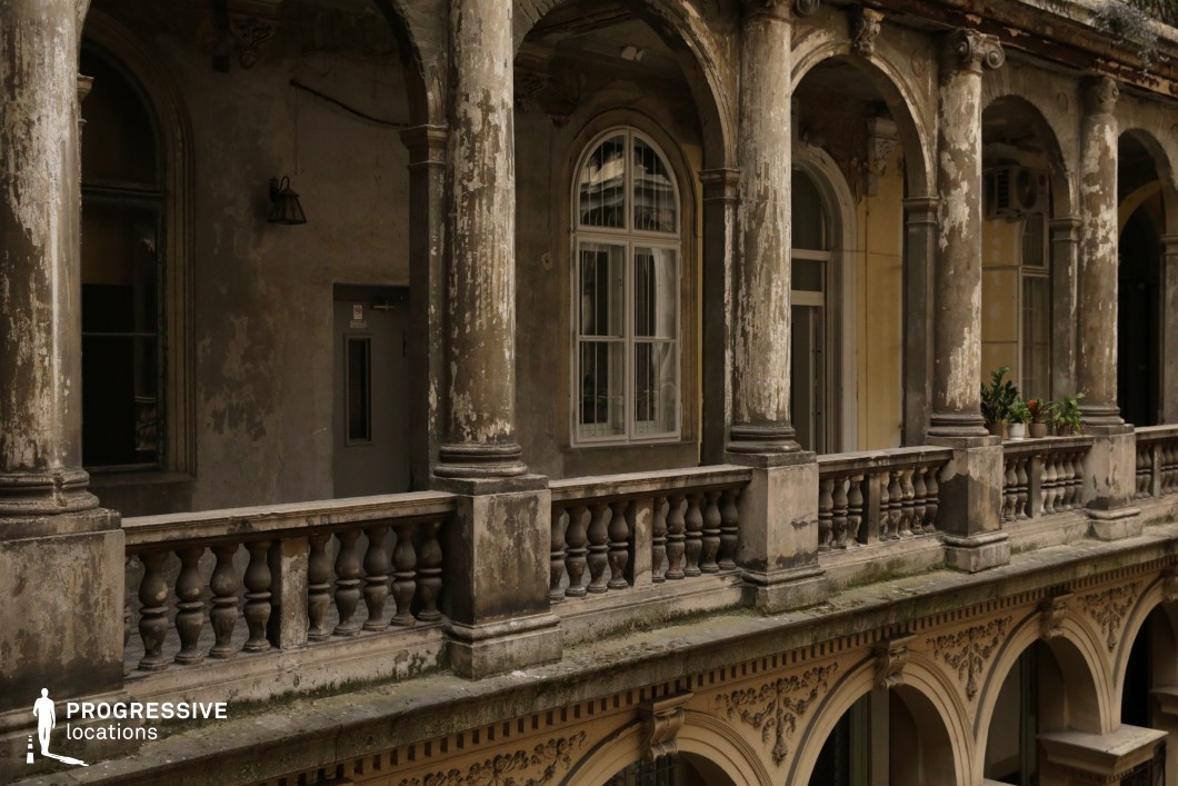 This image depicts an aged, classical-style balcony with ornate arches and fluted columns that show significant weathering. The balustrades, featuring rounded stone spindles, add to the historic charm, while the peeling plaster and discoloration on the walls convey a sense of abandonment or long-term neglect. The mix of architectural elements, including arched windows and decorative moldings, suggests a once-grand residential or institutional building. This setting is well-suited for period dramas, mystery thrillers, or urban decay narratives, offering a visually rich and textured environment that enhances themes of nostalgia, secrecy, or societal decline.