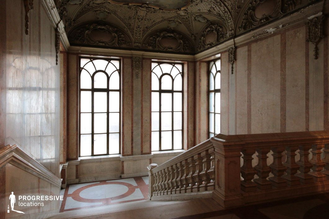 This image showcases a grand staircase in a historic palace, featuring elegant balustrades, high arched windows, and intricate ceiling moldings. The polished marble surfaces, adorned with soft pink and cream tones, reflect the ambient light, creating a luminous atmosphere. The detailed stucco ceiling, embellished with delicate relief work, enhances the regal quality of the space. The large windows introduce a subtle contrast between the bright exterior and the richly textured interior. This location is perfect for filming historical dramas, aristocratic intrigue, period thrillers, or fantasy films, offering a refined and visually striking setting with timeless elegance.