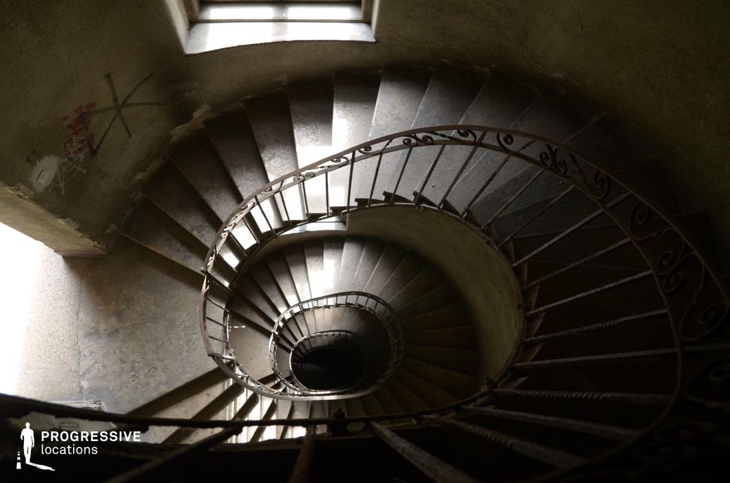 This image showcases a steep, aged spiral staircase with an intricate wrought-iron railing, winding downwards into darkness. The curved structure creates a hypnotic visual effect, emphasized by the interplay of light and shadow filtering through a nearby window. The surrounding walls display signs of wear, including peeling paint and graffiti, reinforcing a sense of abandonment and mystery. The perspective from above heightens the dramatic and disorienting effect of the spiral descent. This location is ideal for filming psychological thrillers, noir films, or horror scenes, providing a visually striking setting for suspenseful sequences, chases, or introspective moments of tension.