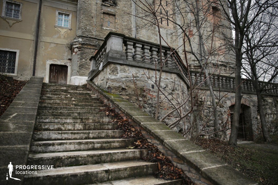This image captures a grand yet decayed stone staircase ascending toward an old building with a deteriorating facade. The balustrades feature classical design elements, though they are visibly worn, with moss and discoloration adding to the aged aesthetic. The structure exhibits a mix of exposed stone and faded plaster, with cracks and missing sections emphasizing its abandoned character. A heavy wooden door, framed by chipped masonry, adds an air of secrecy, while the arched openings and barred windows on the lower level reinforce a sense of history and mystery. This location is ideal for filming gothic horror, period dramas, or historical thrillers, serving as a perfect setting for eerie encounters, hidden secrets, or scenes of suspense.