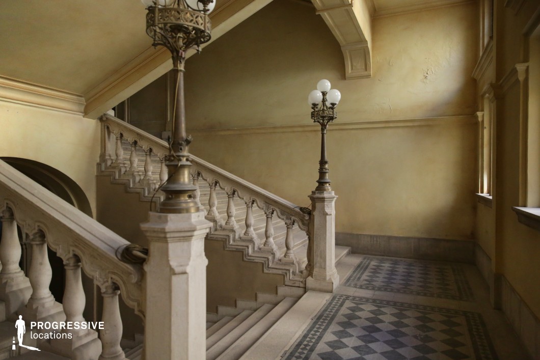 This image captures a historic staircase with intricately carved stone balustrades and elegant wrought-iron street lamps. The soft yellow-hued walls and geometric-patterned tiled flooring create a refined yet slightly aged ambiance. The sweeping stairway, framed by arched openings and high ceilings, enhances the grandeur of the space. The lighting fixtures, featuring detailed metalwork and globe-shaped glass, add a touch of classic sophistication. This location is ideal for period dramas, aristocratic narratives, mystery thrillers, or noir-style films, offering a stately and atmospheric backdrop with a timeless elegance.