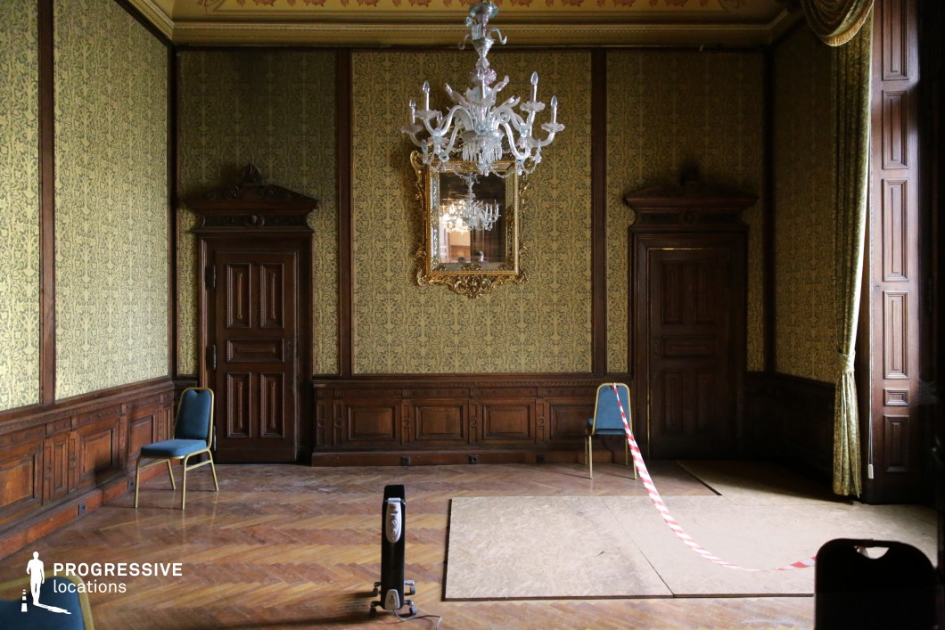 This elegant yet slightly worn interior features rich wood paneling, intricate damask-patterned wallpaper, and a grand chandelier suspended from a decorated ceiling. The symmetrical layout, with two wooden doors flanking a gilded mirror, enhances the sense of balance and classical refinement. The polished parquet flooring adds warmth, while the aged elements, such as faded wallpaper and minor signs of disrepair, introduce a subtle atmosphere of history and nostalgia. The soft lighting and muted tones create a dignified but slightly eerie ambiance. This location is ideal for filming period dramas, psychological thrillers, or gothic mysteries, offering a setting that conveys both sophistication and underlying tension.