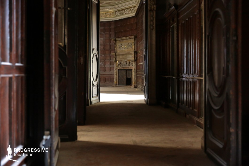 This image presents a dimly lit corridor with rich, dark wood-paneled walls and large, heavy doors leading into an opulent room. The focal point is a grand fireplace adorned with intricate gold detailing, framed by more wood paneling and a coffered ceiling. The long shadows and warm glow from the distant light enhance the dramatic and mysterious atmosphere. The setting exudes an aristocratic, old-world charm, making it an excellent filming location for period dramas, gothic horror, mystery thrillers, or historical intrigue, where an air of secrecy or grandeur is essential to the visual narrative.