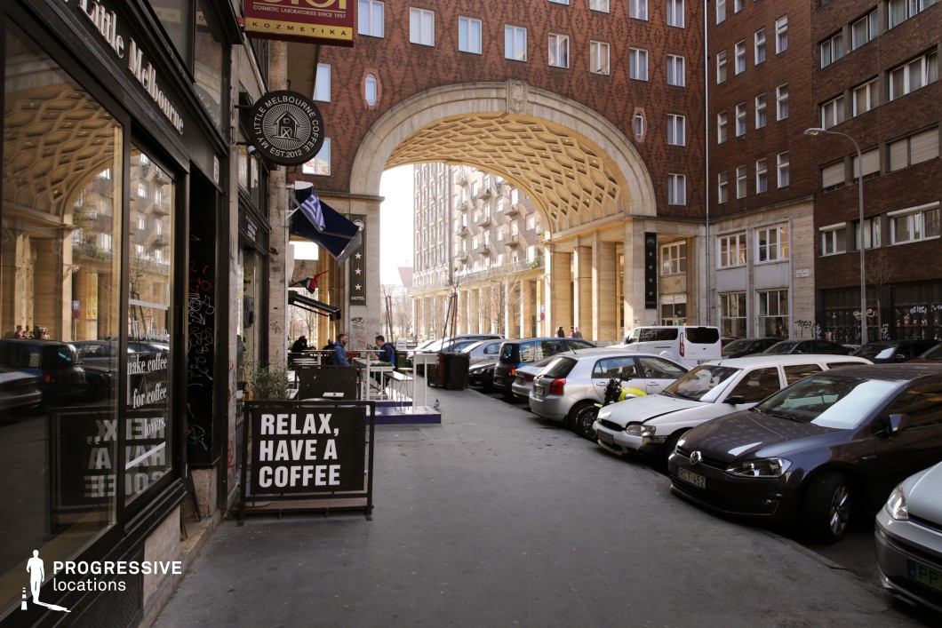 This image captures a dynamic European city street, defined by a monumental brick-clad archway with intricate geometric detailing on its underside. The surrounding architecture blends mid-century modern and classical influences, with large windows and strong vertical elements. On one side, a café with reflective windows and an inviting outdoor seating area adds to the lively street ambiance, while parked cars line the curb, emphasizing the urban setting. The perspective through the archway leads towards another columned building in the background, enhancing depth. This location is ideal for contemporary city dramas, urban exploration narratives, or stylish commercial shoots, offering a visually rich and cinematic backdrop with a blend of historic and modern urban energy.