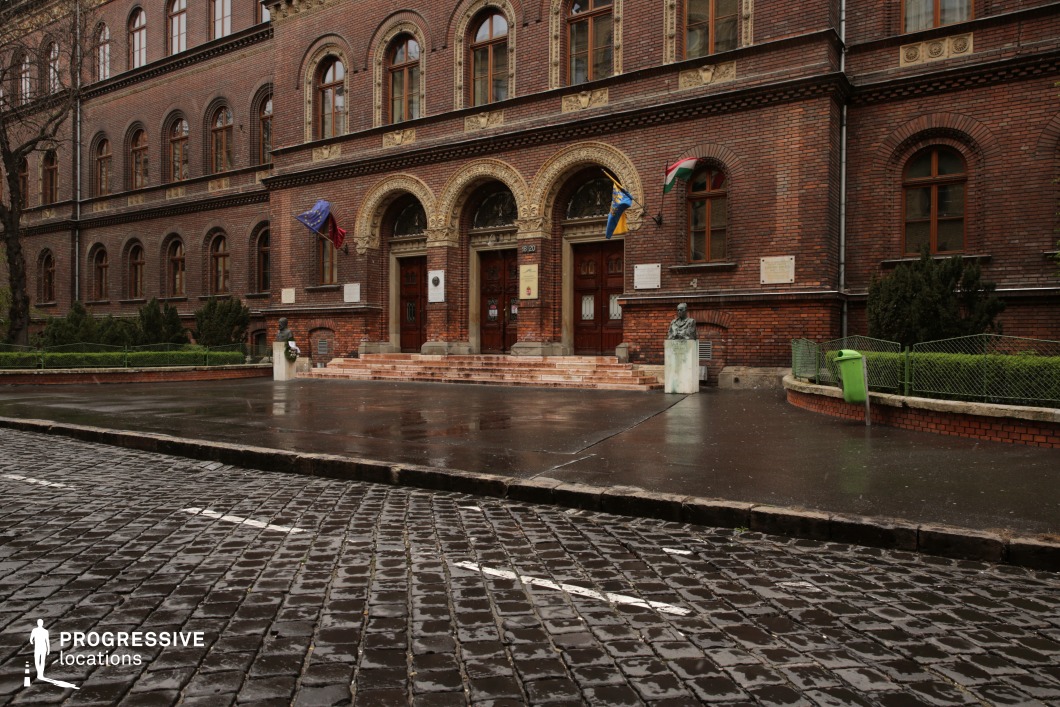 This image features an imposing historic brick building with intricate architectural details. The arched entranceways, adorned with decorative stonework, lead to large wooden doors, adding a sense of grandeur and institutional significance. The facade is characterized by warm red brick with contrasting ornate elements around the windows and entryways. A cobblestone street in the foreground enhances the old-world atmosphere, while a small landscaped area with greenery and statues adds depth and texture. This location is ideal for period dramas, academic or governmental settings, and historical reconstructions, providing a dignified and visually rich backdrop with European architectural heritage.
