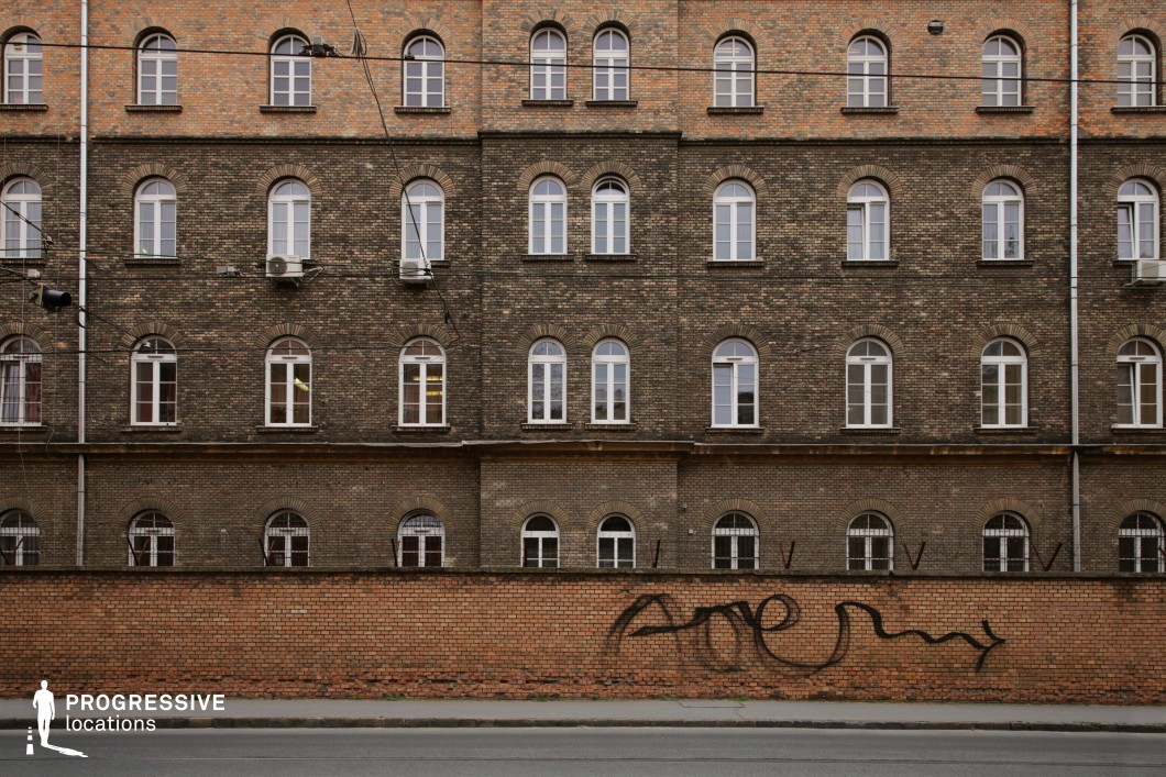 This imposing brick building features a repetitive pattern of arched windows, contributing to its historic and institutional aesthetic. The facade showcases a gradient of aged brick tones, with darker sections adding depth and character. A secondary brick wall in the foreground, marked with graffiti, introduces an urban contrast, while metal spikes atop the wall hint at restricted access. Overhead wires crisscross the composition, enhancing the gritty, structured look. This location is ideal for filming scenes that require an austere, institutional, or even mysterious atmosphere, making it suitable for crime dramas, historical reconstructions, or dystopian settings.