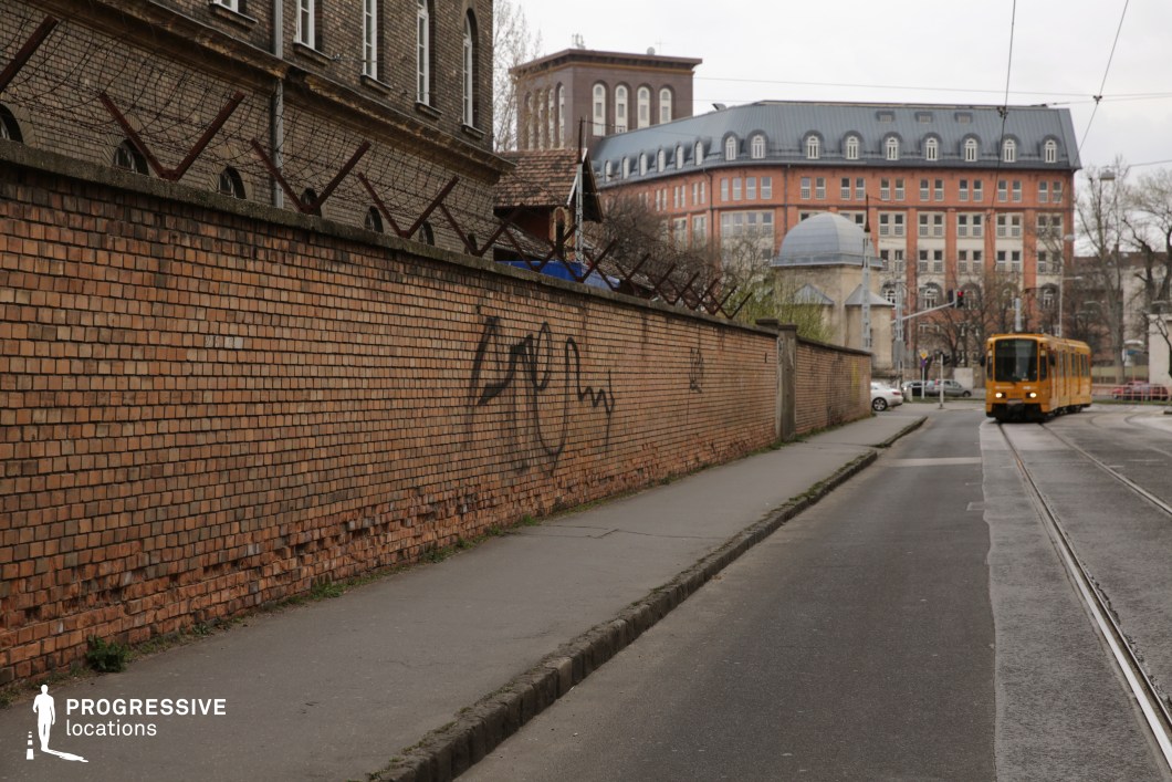 This urban streetscape features a long, aged brick wall topped with barbed wire, giving it a fortified and restrictive appearance. The weathered facade, with visible damage and graffiti, contributes to a gritty, institutional aesthetic. Beyond the wall, a mix of historic and modern buildings with mansard roofs and large windows adds depth and contrast to the setting. A yellow tram curves along the tracks, reinforcing the city’s transit-oriented character and lending a dynamic element to the otherwise enclosed scene. The location’s blend of security features, architectural contrasts, and urban infrastructure makes it ideal for crime dramas, social realism films, or dystopian thrillers, where themes of control, surveillance, or escape are central to the narrative.