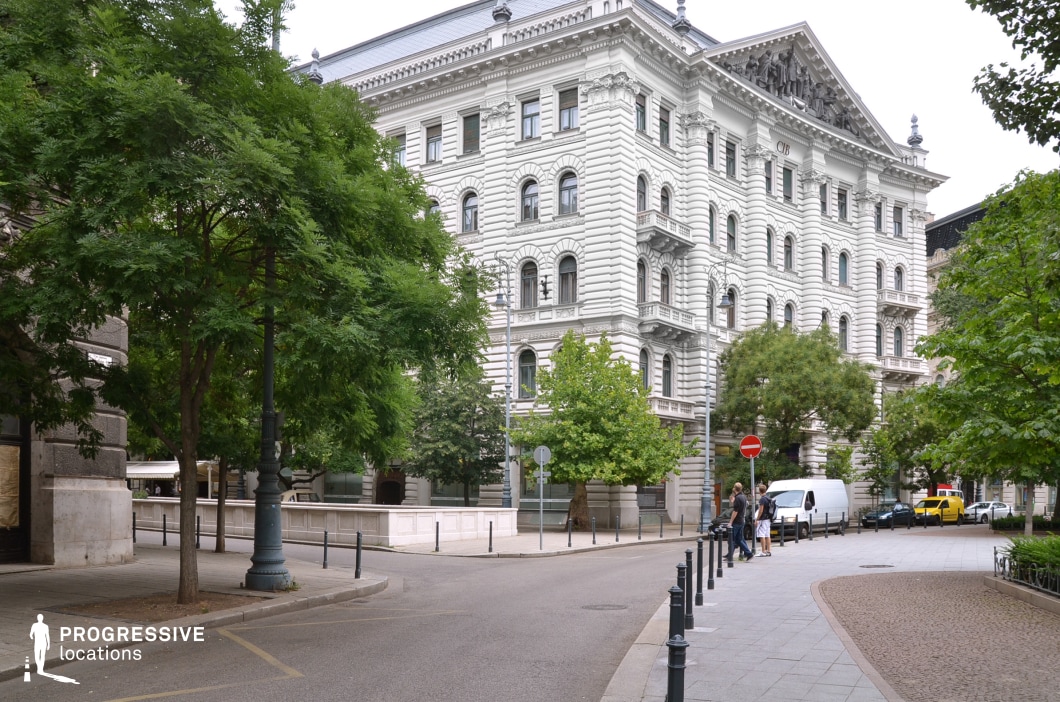 This image captures an elegant European streetscape dominated by a stately white neo-Renaissance building with intricate architectural detailing. The symmetrical facade features ornate pilasters, sculpted reliefs, arched windows, and a grand triangular pediment adorned with classical figures, reinforcing its historic and institutional character. The surrounding area is lined with lush green trees, decorative street lamps, and a gently curving road, creating a refined and picturesque setting. This location is ideal for period films, political dramas, or high-society narratives, offering a sophisticated and cinematic backdrop that blends architectural grandeur with urban greenery.