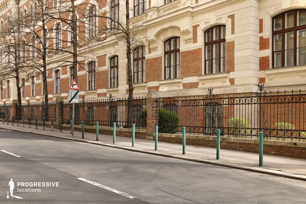 This image showcases a grand historic building with a striking red-brick and cream-colored facade, embodying classic European architectural refinement. The symmetrical windows, framed by decorative stone elements, contribute to its stately appearance, while the wrought-iron fence with intricate patterns adds a sense of exclusivity and prestige. A row of evenly spaced green bollards lines the sidewalk, reinforcing the structured urban layout. The soft contrast between the structured architectural elements and the greenery within the fenced area enhances the setting's visual appeal. This location is ideal for academic or governmental scenes, period dramas, or high-society narratives, offering a distinguished and formal backdrop with a refined atmosphere.