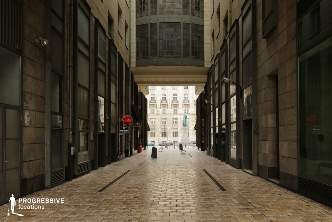 This image features a covered passageway with a striking blend of modernist and classical architectural elements. The corridor is flanked by tall, dark stone-clad walls with large industrial-style windows, creating a structured, almost austere ambiance. Overhead, a distinctive Art Nouveau-style bay window protrudes, adding a decorative yet imposing architectural focal point. The passageway opens up to a grand European square, where a historic stone-clad building and a central statue enhance the sense of depth and contrast. This location is ideal for noir thrillers, espionage films, or dramatic urban storytelling, offering a visually dynamic and slightly enigmatic setting.