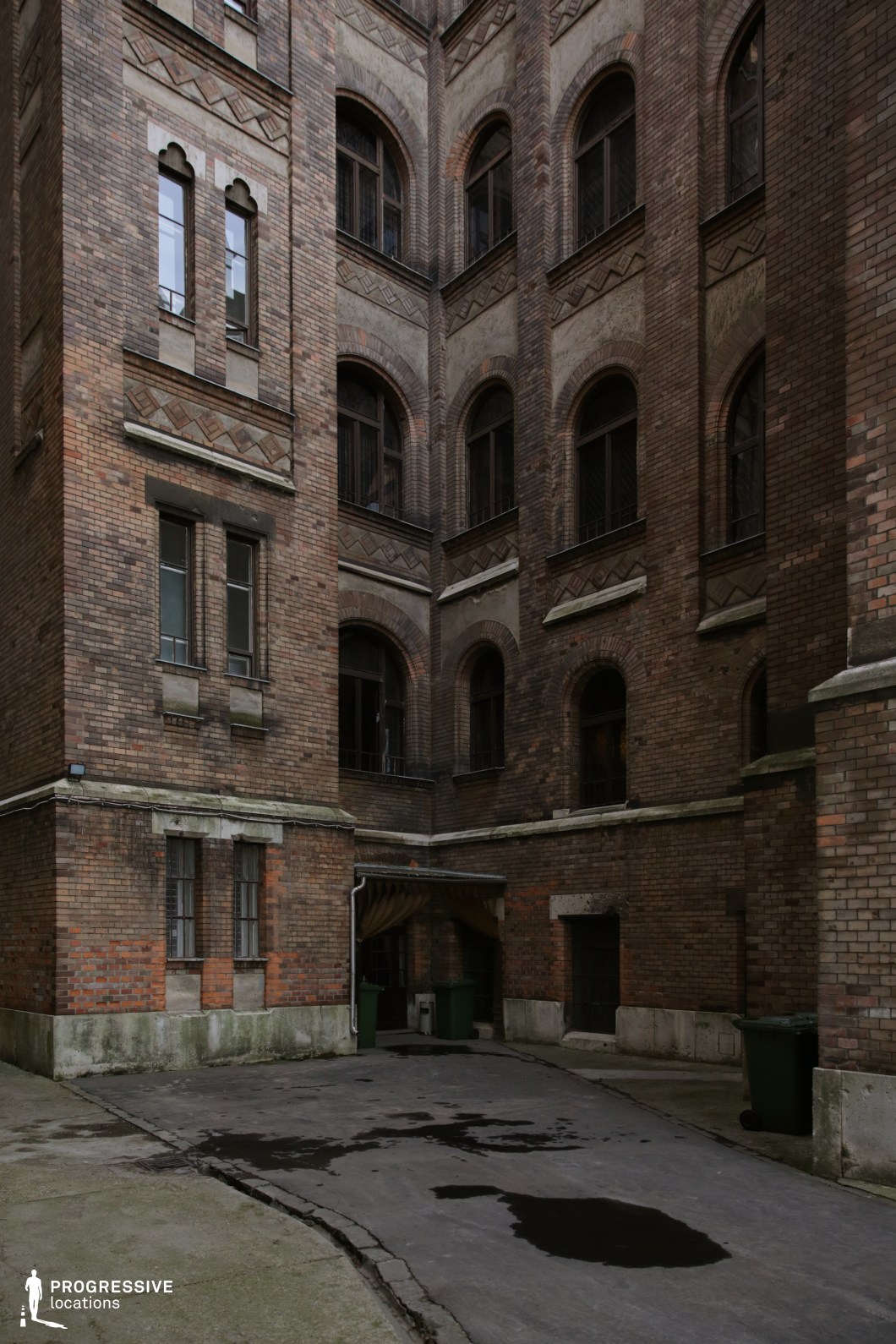 This enclosed courtyard is surrounded by towering brick facades with arched windows, featuring intricate decorative brickwork that hints at a late 19th or early 20th-century institutional or residential complex. The aged walls, darkened by time, create a sense of history and seclusion, while the narrow, windowed openings and barred lower windows reinforce an air of control and restriction. The worn pavement, scattered stains, and minimal lighting contribute to an eerie, almost foreboding ambiance. The confined space, high walls, and lack of open exits make this location highly suitable for psychological thrillers, period dramas with a melancholic tone, or crime narratives involving hidden secrets and confined spaces.