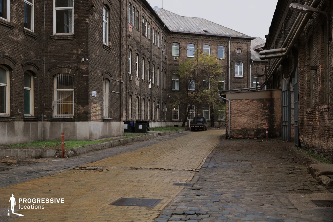 This enclosed courtyard is framed by dark, weathered brick buildings featuring arched windows, some secured with metal bars, reinforcing an institutional or industrial past. The cracked and uneven yellow cobblestone pathway contrasts with the surrounding damp, darkened stone, adding a sense of history and wear. Overhead pipes and exposed utility elements further contribute to the utilitarian aesthetic, while patches of grass and a lone tree introduce subtle natural elements. The enclosed nature of the space, along with its aged textures, makes it an excellent setting for gritty crime dramas, historical narratives, or urban thrillers, offering a raw, atmospheric backdrop for stories of secrecy, survival, or forgotten pasts.