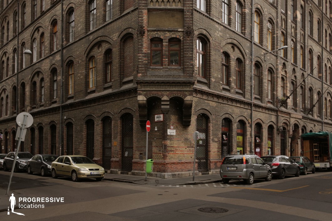 This image captures a striking corner building with a dark brick facade, featuring arched windows and decorative masonry that evoke late 19th-century European architecture. The structure has a slightly weathered appearance, with variations in brick tones adding to its aged character. The ground floor features a series of closed, gated archways, hinting at a past commercial or industrial function. The narrow street, lined with parked cars, enhances the enclosed urban atmosphere, while the muted lighting and subdued color palette contribute to a moody, cinematic quality. This location is ideal for noir thrillers, historical dramas, or crime films, offering an evocative and textured setting with a sense of intrigue and history.