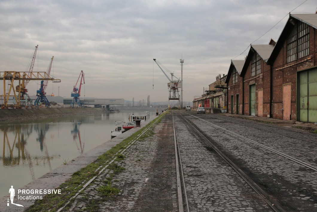 This expansive dockside location features a long row of aged brick warehouses with large metal doors, set against a cobblestone-paved rail track running parallel to the calm waters of the river. Towering industrial cranes in various colors line the opposite bank, emphasizing the port’s utilitarian nature. The overgrown patches of grass, rusting metal fixtures, and weathered facades contribute to an atmosphere of abandonment or industrial decline. The long, uninterrupted sightlines and mix of heavy infrastructure make this setting ideal for crime thrillers, neo-noir films, or action sequences involving smuggling, covert meetings, or high-stakes pursuits. The open expanse and gritty textures also suit dystopian and post-industrial storytelling.