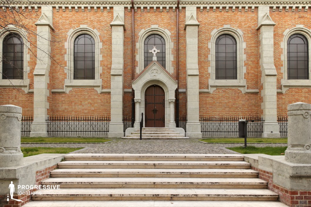 This impressive church facade features a symmetrical design with arched windows, strong vertical stone elements, and a grand entrance framed by an ornate pediment. The red brick walls contrast with the light-colored stone details, emphasizing the Gothic architectural influence. A set of wide, weathered steps leads up to the entrance, adding depth and formality to the composition. The black wrought iron fence encircling the churchyard reinforces a sense of enclosure and reverence. This location is ideal for filming historical dramas, religious narratives, or suspenseful mystery scenes, offering a dignified and atmospheric setting.