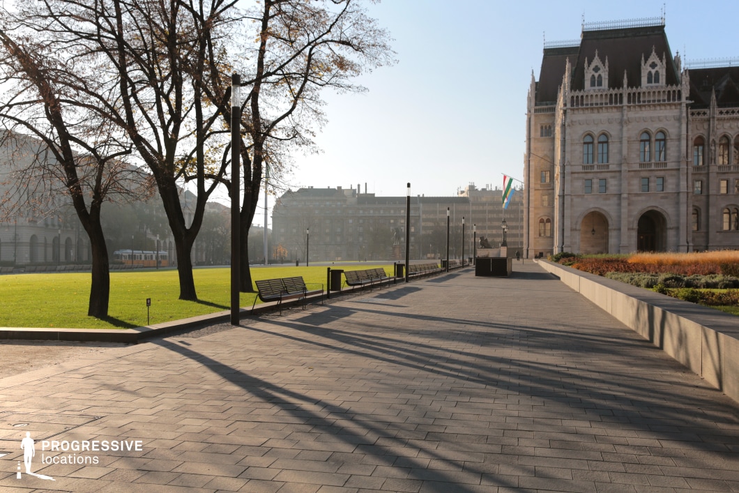 This expansive public square blends historical elegance with modern urban design. A grand neo-Gothic building with intricate detailing and pointed rooftops dominates the right side, exuding an air of prestige and formality. The foreground features a wide, paved pedestrian walkway lined with benches, street lamps, and neatly landscaped greenery, leading toward an open green lawn framed by mature trees. In the background, a mix of classical and modern buildings adds depth and contrast to the cityscape. This location is ideal for filming political dramas, historical narratives, or contemplative scenes, offering a majestic yet open setting that balances grandeur with public accessibility.