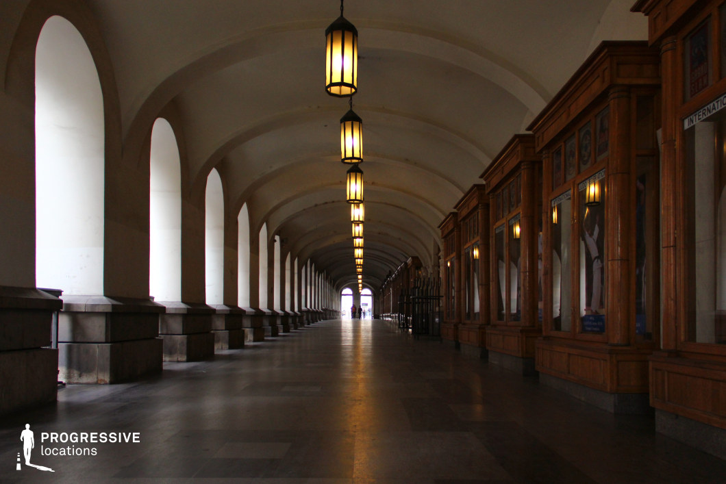 This grand, covered arcade features a rhythmic sequence of tall, rounded arches supported by sturdy stone bases, creating a sense of depth and perspective. Hanging lanterns illuminate the passageway, casting a warm glow that enhances the corridor’s historic character. On one side, a series of wooden-framed display windows with intricate detailing add a refined, old-world charm, suggesting a cultural or commercial function. The high, vaulted ceiling and long, uninterrupted sightline contribute to an atmospheric and cinematic quality. This location is ideal for period dramas, mystery thrillers, or elegant European cityscapes, offering a setting for scenes requiring architectural grandeur, dramatic lighting, or a sense of quiet intrigue.