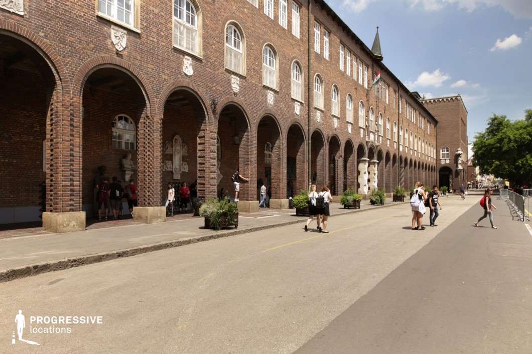This striking historic building features a long row of evenly spaced brick archways, creating a strong sense of rhythm and depth. The rich brown brick facade is adorned with decorative stone reliefs and arched windows, blending Romanesque and early 20th-century institutional architectural styles. The covered walkway beneath the arches adds a dramatic visual element, leading toward an open plaza and tree-lined street. The structured elegance of the location makes it ideal for filming academic, historical, or cultural narratives, as well as atmospheric scenes for period dramas or coming-of-age stories set in a European cityscape.