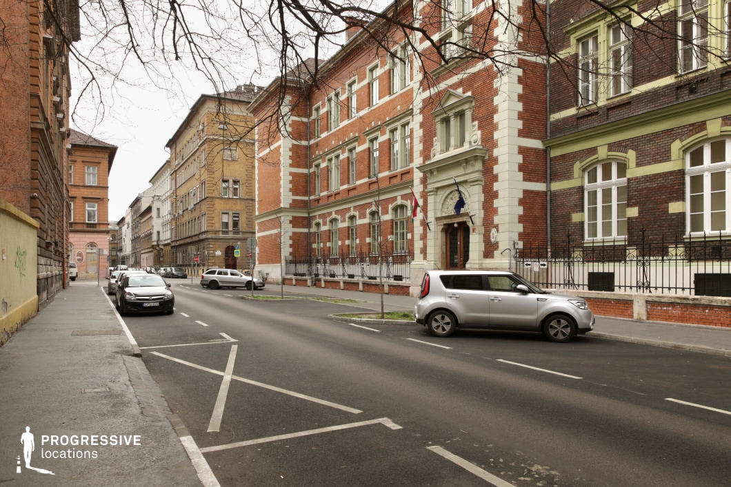 The image captures a European-style urban street lined with historic brick buildings, showcasing a mix of late 19th and early 20th-century architectural styles. The prominent red brick structures feature decorative white stone accents around windows and doorways, intricate wrought-iron fencing, and classical detailing on entrances. The street itself is relatively narrow, flanked by a sidewalk and a row of parked vehicles, with a clear vanishing point leading into the background. The muted color palette of beige, red, and dark metal railings gives the scene a subdued yet elegant character. The location is ideal for period films, crime dramas, or European-set political thrillers, offering a sophisticated yet slightly somber atmosphere suitable for formal or institutional settings.