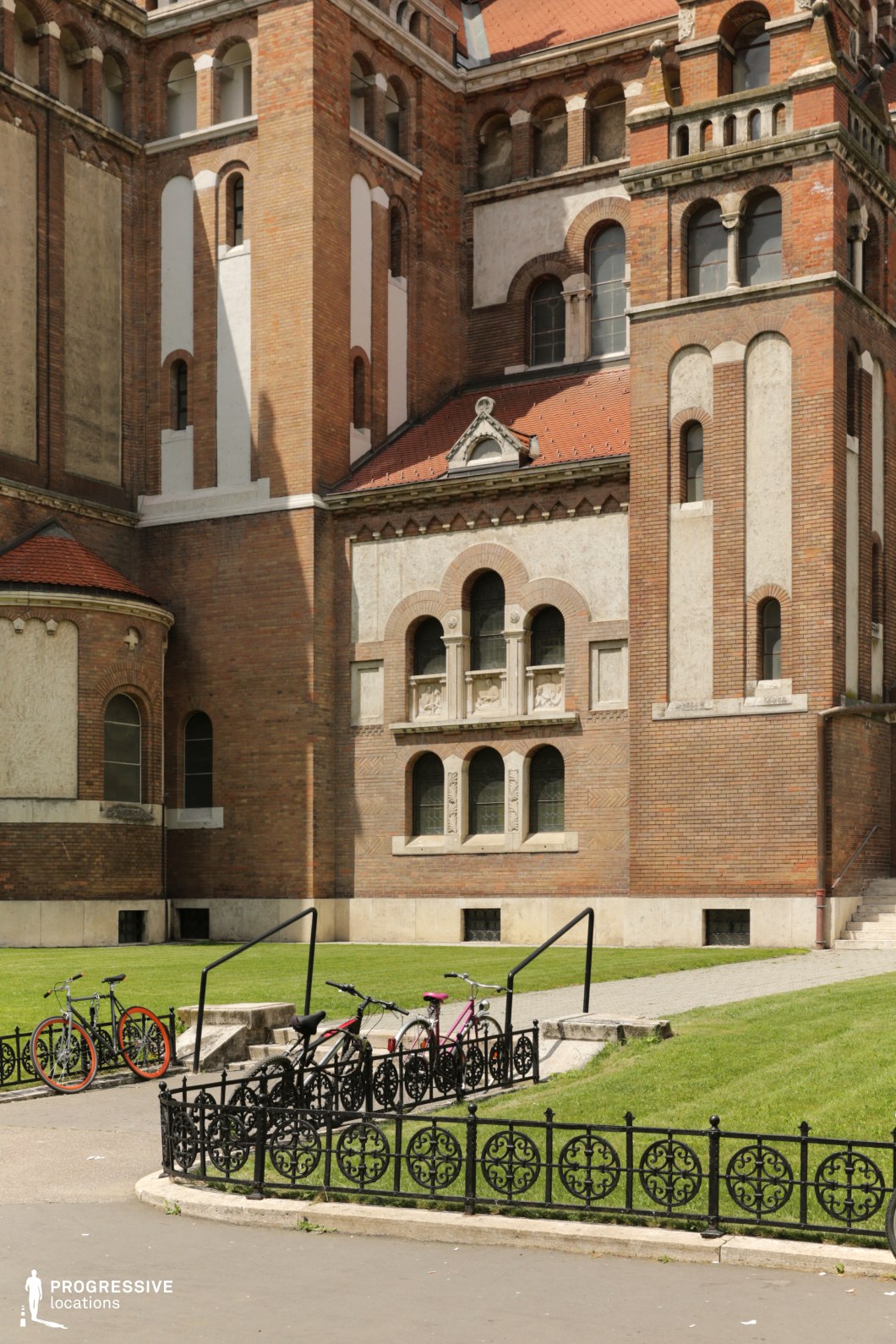 This historic brick building showcases a striking mix of Romanesque and Gothic architectural elements, with arched windows, intricate stone detailing, and decorative cornices. The reddish-brown brick facade is accented with contrasting lighter stonework, enhancing the depth and texture of the structure. The foreground features a manicured green lawn, a paved walkway, and an ornate wrought iron fence, contributing to the refined and structured aesthetic. This location is ideal for filming period dramas, historical documentaries, or mystery-themed productions, offering a grand yet atmospheric setting with rich architectural detail.