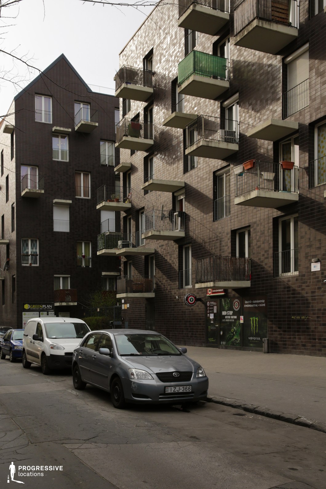 This image presents a contemporary residential street dominated by modern apartment buildings with dark brick facades and a striking pattern of protruding balconies. The repetitive geometric design of the balconies, featuring a mix of metal railings, glass panels, and wooden elements, adds depth and texture to the scene. The lower level of the building houses small commercial spaces, subtly integrated into the structure. The subdued color palette and minimalist design give the area a functional, slightly austere atmosphere. This location is well-suited for urban dramas, social realism, or crime thrillers, offering an authentic, modern European residential backdrop with a sense of everyday life and quiet tension.