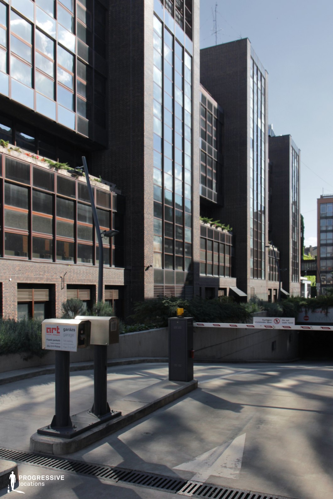 This urban location features a striking modernist office complex with a dark brick facade and large reflective glass windows that create a sleek, corporate aesthetic. The geometric repetition of windows and vertical lines enhances the contemporary architectural style. Lush greenery integrated into the building’s design adds a touch of organic contrast. In the foreground, an underground parking entrance with a barrier and security system introduces a functional element, reinforcing the setting's metropolitan nature. This location is ideal for filming corporate dramas, crime thrillers, or espionage sequences, providing a dynamic urban backdrop with a mix of structured modernism and subtle Brutalist influences.
