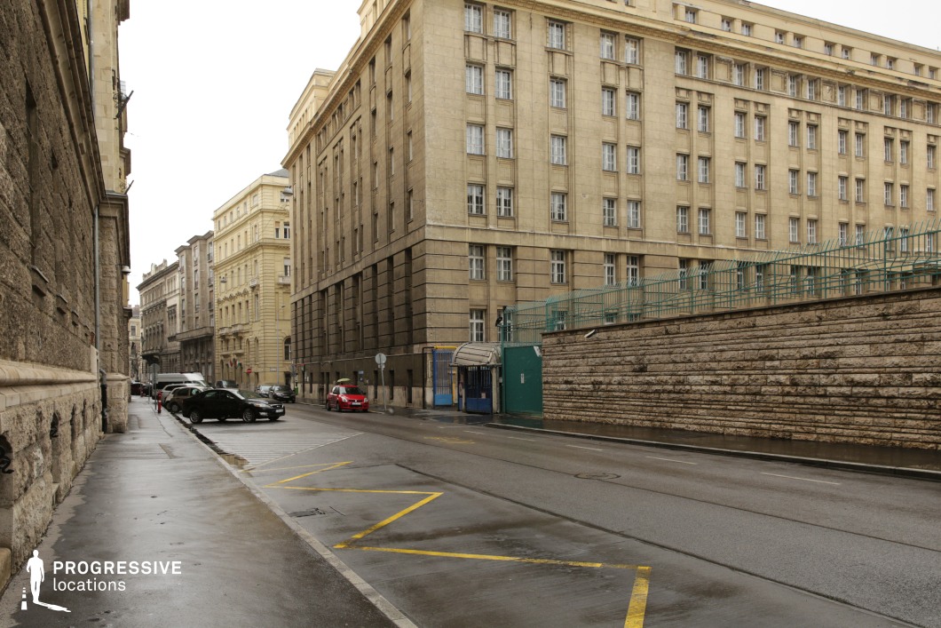 This imposing institutional-style building dominates the scene with its rigid, symmetrical facade and repetitive window patterns, evoking mid-20th-century government or administrative architecture. The beige stone construction contrasts with the textured retaining wall running parallel to the street, which is topped with metal fencing, adding an element of security and control. The street is relatively wide, with marked parking spaces and a mix of historic and utilitarian structures in the background, providing a layered urban setting. The austere and somewhat sterile atmosphere of this location makes it ideal for crime dramas, political thrillers, or scenes requiring a bureaucratic, official, or high-security environment. The combination of strong architectural lines and muted tones also lends itself well to dystopian or espionage-themed productions.