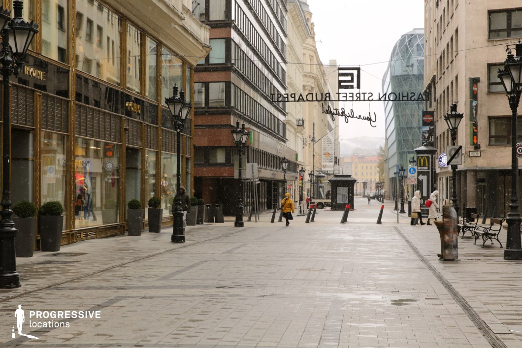 This pedestrianized street showcases a blend of historic and contemporary architecture, creating a visually dynamic urban space. On the left, an ornate building with gold-trimmed glass storefronts exudes a sense of classic luxury, while the right side features a mix of stone-facade structures and sleek modern glass constructions. Decorative street lamps line the walkway, reinforcing a European-style ambiance. The paved street, devoid of vehicular traffic, offers an open and inviting perspective leading towards a background of more historic buildings and a glass-enclosed structure. This location is ideal for filming elegant urban sequences, commercial shoots, or dramatic scenes that require a blend of classic and modern city aesthetics. The setting suits genres like romance, drama, and upscale fashion commercials.