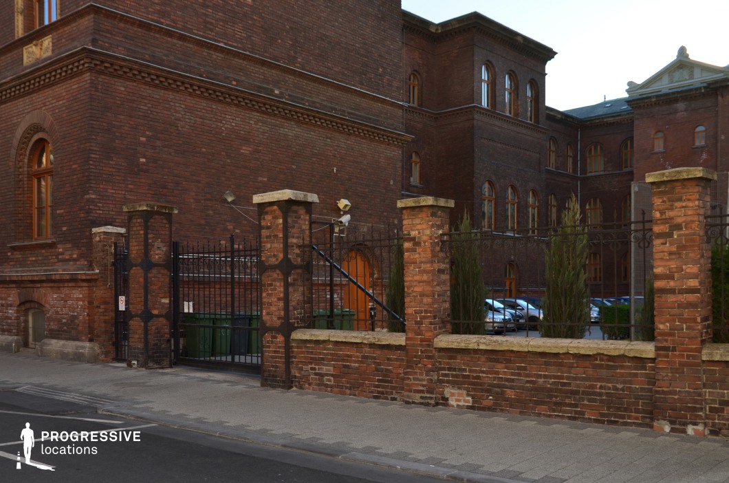 This imposing red-brick building features a strong institutional character, with arched windows, decorative brickwork, and a structured, symmetrical facade. The surrounding iron-fenced courtyard, framed by weathered brick columns, adds a sense of enclosure and formality. The aged masonry and dark wrought iron gate enhance the location's historical depth, suggesting a prestigious or authoritative setting. This location is ideal for filming period dramas, academic or governmental narratives, or mystery and crime scenes, offering a visually rich and atmospheric backdrop with a sense of history and security.