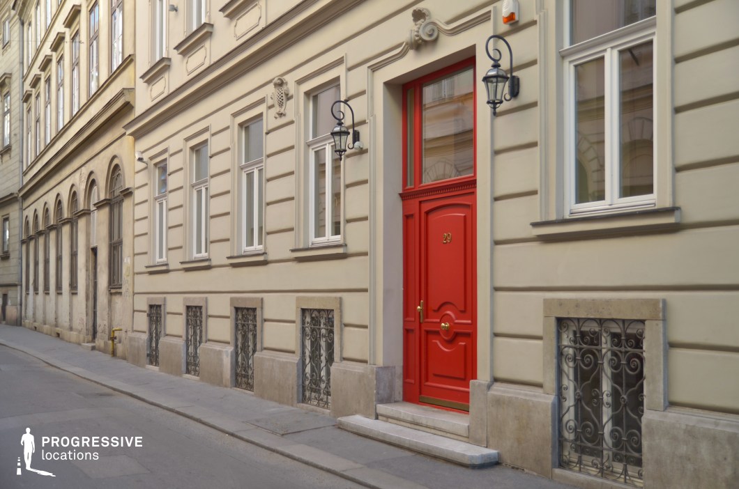 This refined historic townhouse facade features a symmetrical design with cream-colored walls, decorative molding, and wrought iron window grilles, exuding a classic European charm. The bold red entrance door, framed by intricate detailing and flanked by two ornate wall-mounted lanterns, acts as a striking focal point. The narrow cobblestone street and aged architectural elements in the background enhance the intimate and timeless atmosphere of the setting. This location is ideal for filming sophisticated period dramas, mystery narratives, or romantic films, providing an elegant yet understated urban backdrop with a distinct old-world character.
