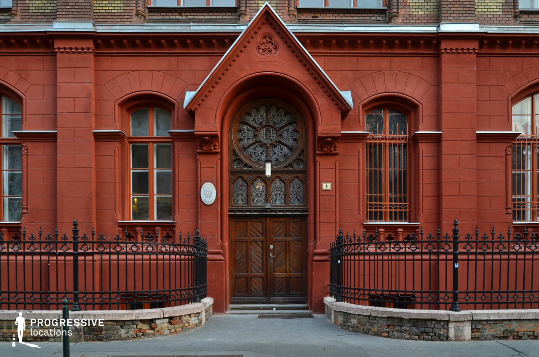 This image showcases a grand red-brick building with a richly decorated entrance, embodying Gothic Revival architectural elements. The arched doorway is framed by intricate stone carvings, while the wooden double doors feature elaborate wrought-iron and stained-glass detailing, adding an air of historic sophistication. The symmetrical façade is punctuated by tall, arched windows with decorative iron grilles, reinforcing the structure's formal and institutional character. A black wrought-iron fence with intricate patterns encloses the entrance area, adding to the building’s distinguished aesthetic. This location is ideal for period dramas, academic or historical settings, and mystery-themed productions, offering a visually compelling and atmospheric backdrop.