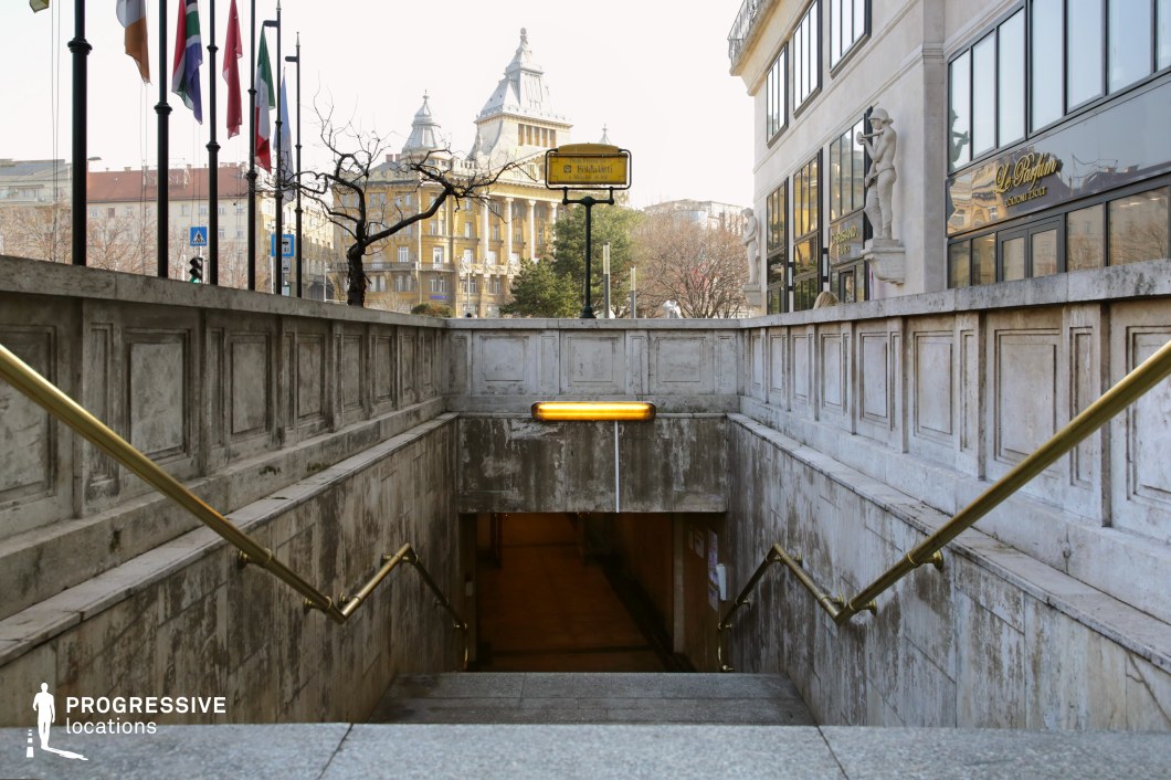 This image captures a descending entrance to an underpass, framed by aged stone walls with decorative paneling and golden metal handrails. The slightly worn texture of the stone surfaces contrasts with the refined architectural details of the surrounding buildings. To the right, a stately structure adorned with sculptural elements and large reflective windows adds a sense of elegance. In the background, a historic building with a distinctive roofline and ornate facade reinforces the classic European cityscape ambiance. This location is ideal for filming urban drama, noir-inspired thrillers, or mystery sequences, offering a visually compelling transition between street-level and underground environments.