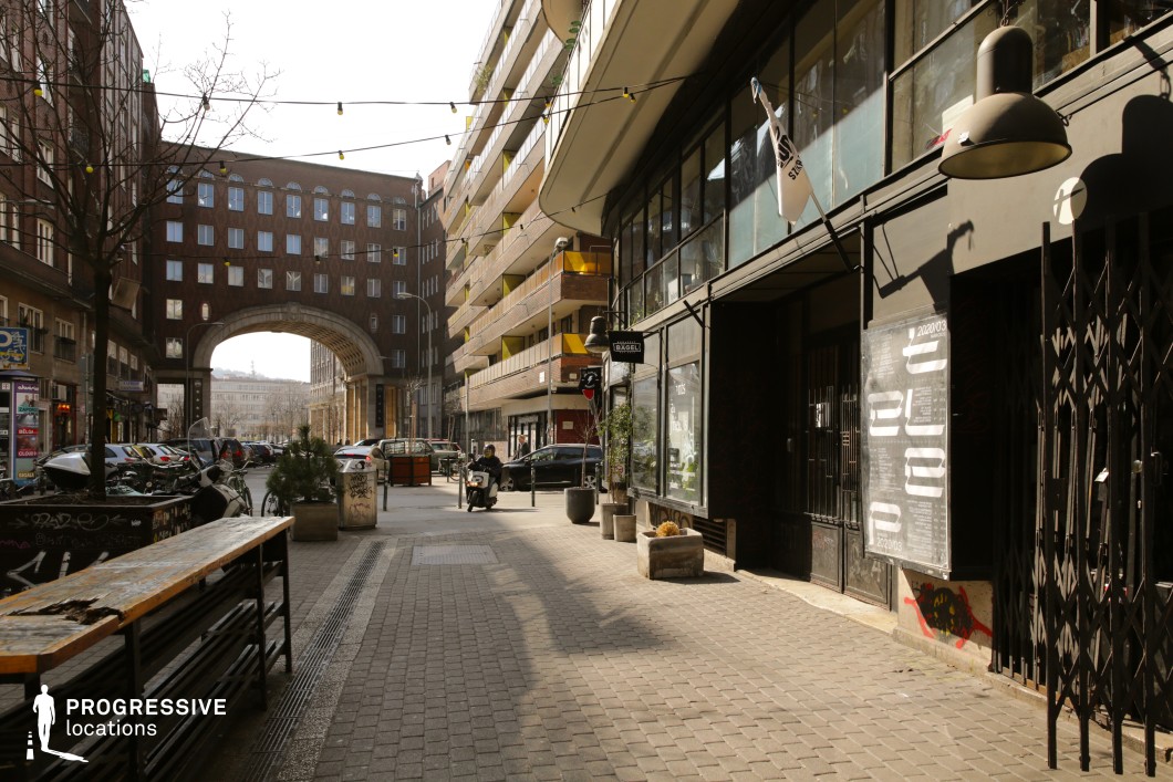 This urban street scene blends historic and contemporary elements, featuring a striking brick archway framing the background. The street is lined with mixed-use buildings, including modern residential balconies with vibrant yellow railings on one side and an industrial-style storefront with large windows and metal grates on the other. String lights above add a touch of atmosphere, while the textured pavement and outdoor seating hint at a vibrant, social setting. The combination of architectural contrast and depth makes this location ideal for filming urban dramas, independent films, or street culture documentaries, offering a dynamic and visually rich city backdrop.