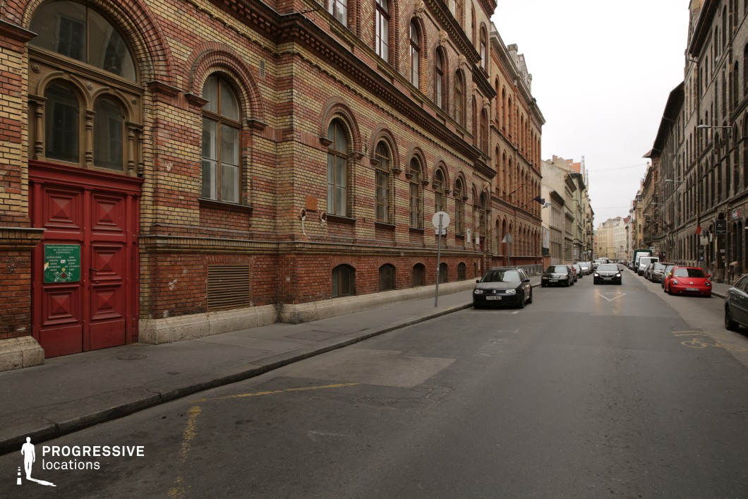 This image captures a narrow European street lined with grand historic brick buildings, exuding late 19th-century architectural elegance. The detailed façades feature arched windows, decorative brick patterns, and ornate cornices, contributing to the rich visual texture. A striking red double-door entrance contrasts with the muted yellow and red brick tones, adding a distinctive focal point. The perspective of the street, flanked by symmetrical rows of buildings, leads toward a vanishing point, enhancing depth. This location is ideal for period dramas, historical reconstructions, or atmospheric urban storytelling, offering a visually cohesive and architecturally rich backdrop.