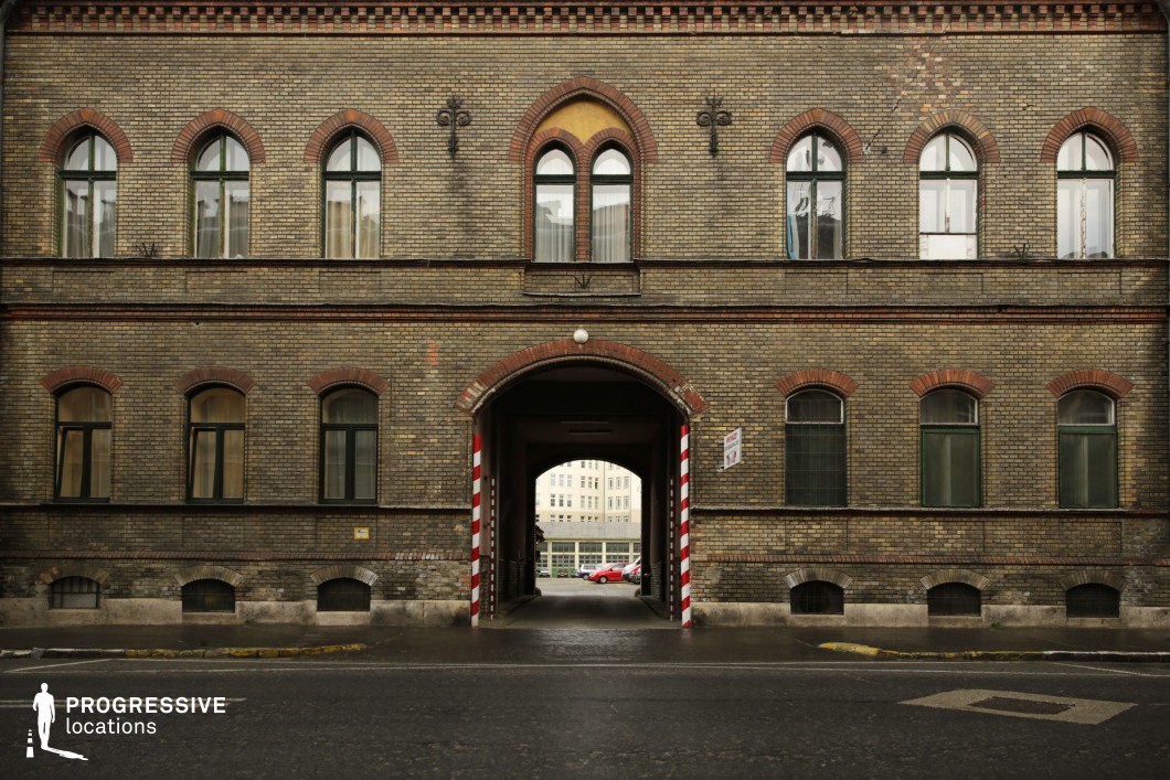 This symmetrical, Victorian-era brick building features arched windows with red brick trim, giving it a distinct industrial or institutional character. The aged yellow and brown brickwork, accented by wrought-iron wall decorations, adds to the building’s historic atmosphere. A central archway, framed with red-and-white striped markings, leads into a courtyard, offering a layered perspective and a sense of controlled access. The contrast between the darkened facade and the bright background visible through the passage enhances the visual depth of the composition. This location is well-suited for period dramas, institutional narratives, or crime thrillers, where themes of authority, secrecy, or urban history play a key role in the visual storytelling.
