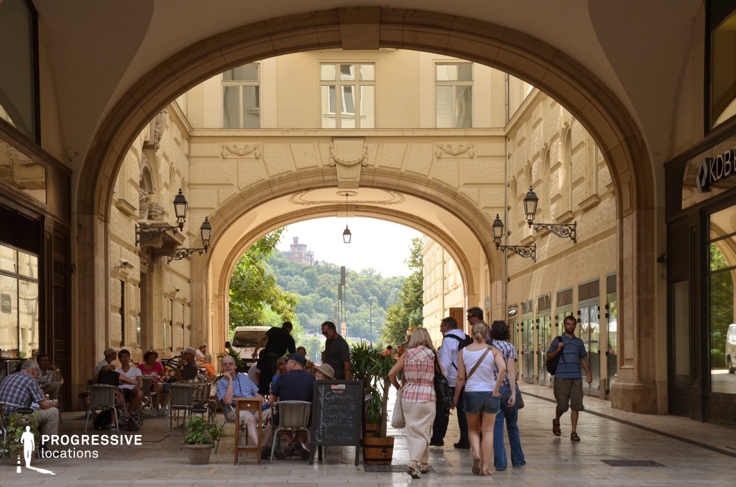 The image captures a beautifully designed archway passage leading into an open area with a lush, green hillside and a historic building in the distance. The architecture features intricate stucco details, wrought-iron lanterns, and large arched openings that frame the inviting outdoor space. The passage is lined with shops and a charming café, where tables and chairs spill into the covered walkway, enhancing the European ambiance. The arch itself serves as a dramatic framing element, drawing the eye toward the picturesque backdrop. This location is ideal for filming urban dramas, romantic encounters, or sophisticated travel-themed narratives, offering a rich blend of historical charm and lively city atmosphere.