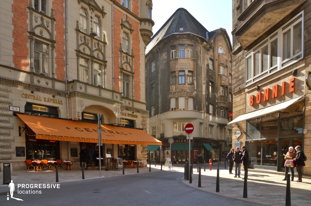 This image captures a European street corner where historic and modern elements blend seamlessly. On one side, a well-preserved, ornate building features a grand café with an inviting orange awning and elegant outdoor seating, exuding a classic charm. In contrast, an aged, weathered structure with peeling plaster and darkened brick stands prominently in the background, creating a striking juxtaposition of old and restored architecture. The street is paved with smooth stone and pedestrian-friendly bollards, leading towards boutique storefronts and a gallery entrance. This location is ideal for filming urban dramas, romantic films, or period pieces, offering a rich and visually compelling city atmosphere.