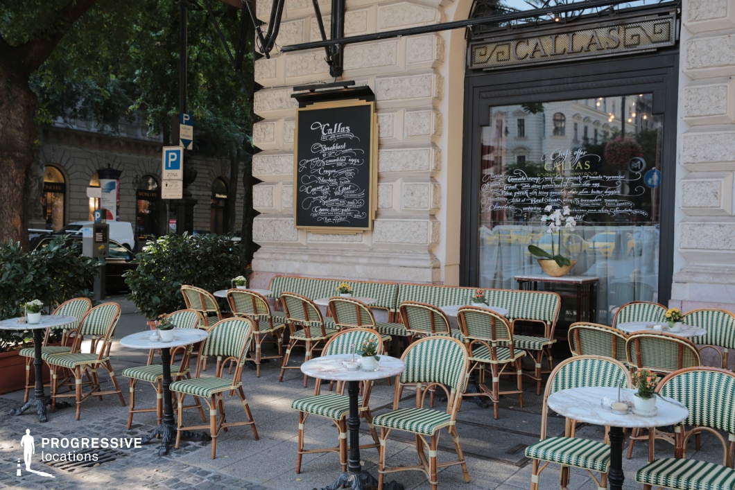 The image showcases a stylish European café terrace set against a historic stone building with intricate detailing. The outdoor seating area features woven rattan chairs and marble-top tables arranged in an inviting manner, creating a quintessential Parisian-style ambiance. A large window with elegant gold lettering and a chalkboard menu enhance the café’s sophisticated aesthetic. Lush greenery and classic lampposts contribute to the refined, cosmopolitan atmosphere. This location is ideal for filming romantic comedies, travel documentaries, or upscale social dramas, offering an effortlessly charming and timeless European setting.