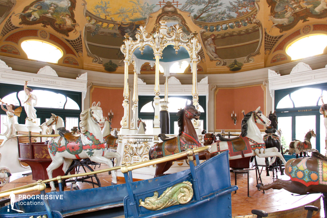 A beautifully preserved vintage carousel housed within an opulent pavilion, featuring intricately carved horses with elaborate saddles, rich colors, and gold accents. The ceiling is adorned with hand-painted murals depicting classical scenes, adding to the nostalgic and elegant atmosphere. Large arched windows framed in deep green allow natural light to highlight the delicate architectural details, while ornamental columns and sculpted angels enhance the fairytale-like setting. This visually striking location is ideal for filming period pieces, fantasy, or whimsical romance, offering a timeless and enchanting backdrop with a dreamlike quality.