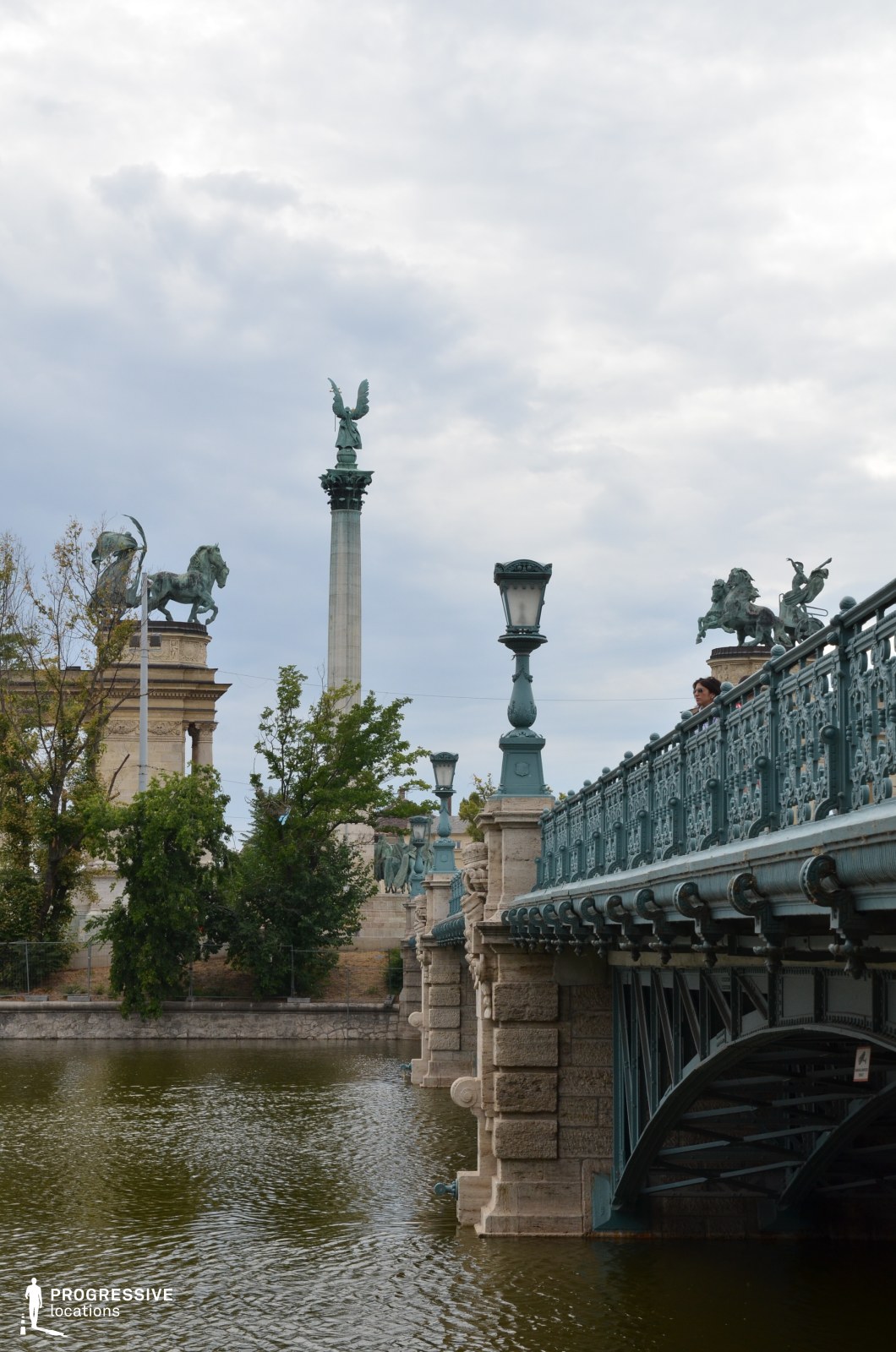 This image showcases an elegant European bridge with intricate cast-iron railings and vintage street lamps, extending over a calm body of water. The bridge’s stone supports and detailed metalwork contribute to its historic character. In the background, grand statues and a towering column adorned with a winged figure create a dramatic and monumental scene. The combination of water, architecture, and sculptural elements offers a strikingly cinematic composition. This location is ideal for filming historical epics, fantasy sequences, or dramatic encounters, providing a regal and timeless setting evocative of classical European grandeur.