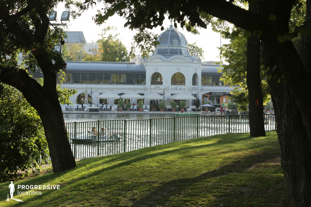 A grand, historic pavilion with elegant domed architecture overlooks a tranquil lake, creating a picturesque contrast between classical design and natural elements. The building’s white façade, arched windows, and decorative metal roofing add sophistication, while the lakeside terrace, adorned with umbrellas and seating, enhances its inviting atmosphere. A gently sloping grassy area in the foreground, framed by mature trees and a metal fence, provides depth and perspective. This location is ideal for filming romantic, historical, or high-society scenes, offering a serene yet refined ambiance reminiscent of European elegance.